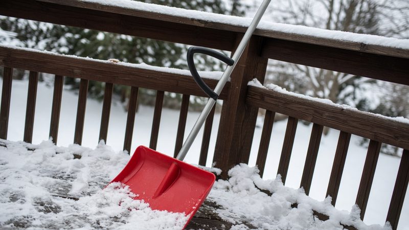 A red snow shovel with a second curved handle leaning on a snowy deck railing, in Waukee, Iowa